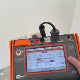 ELEKTRO-TOM - Miernik Sonel MPI-536 podłączony do gniazdka elektrycznego podczas wykonywania pomiarów rezystancji izolacji, widoczny postęp pomiaru na ekranie urządzenia, w tle biała ściana i fragment podłogi...