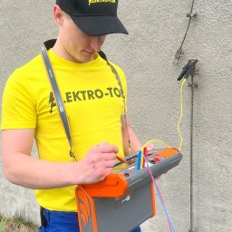 ELEKTRO-TOM - Elektryk w żółtej koszulce i czapce Elektro-Tom wykonuje pomiary uziomu za pomocą miernika, widoczne przewody i uziom przy ścianie budynku.