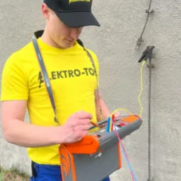 Elektryk w żółtej koszulce i czapce Elektro-Tom wykonuje pomiary uziomu za pomocą miernika, widoczne przewody i uziom przy ścianie budynku.