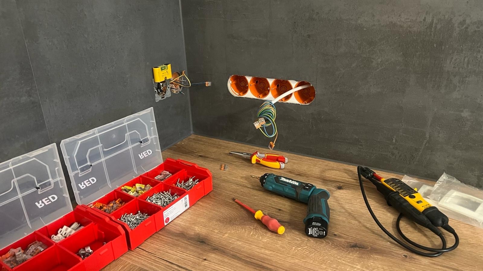 Montaż gniazdek elektrycznych w szarej ścianie, widoczne narzędzia: wkrętarka Makita, tester napięcia Fluke T150, skrzynka z elementami montażowymi BED.