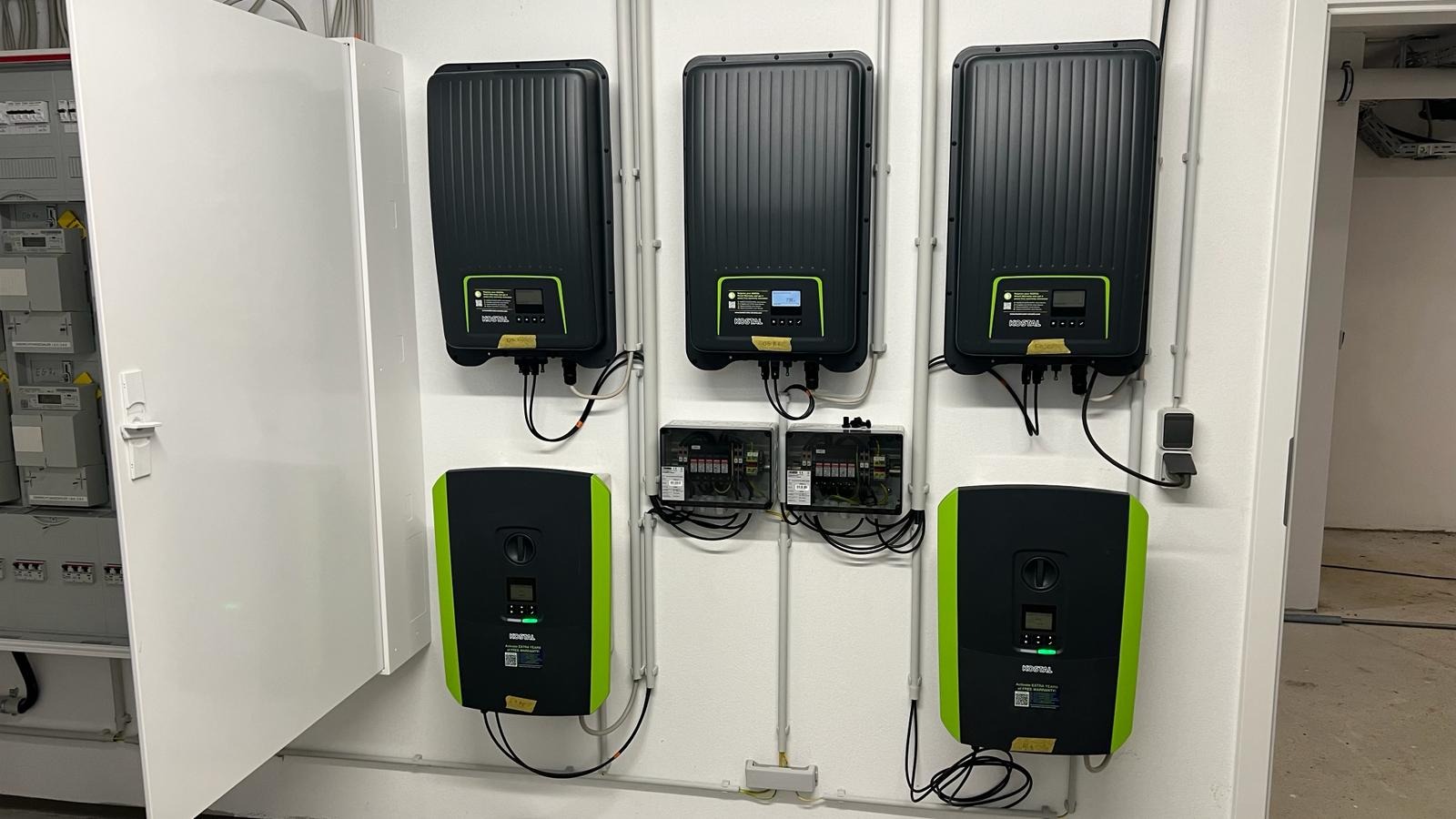 Szereg falowników fotowoltaicznych marki Kostal zamontowanych na białej ścianie, połączonych przewodami w rurkach PCV, z widoczną rozdzielnicą elektryczną po lewej stronie i gniazdkami...
