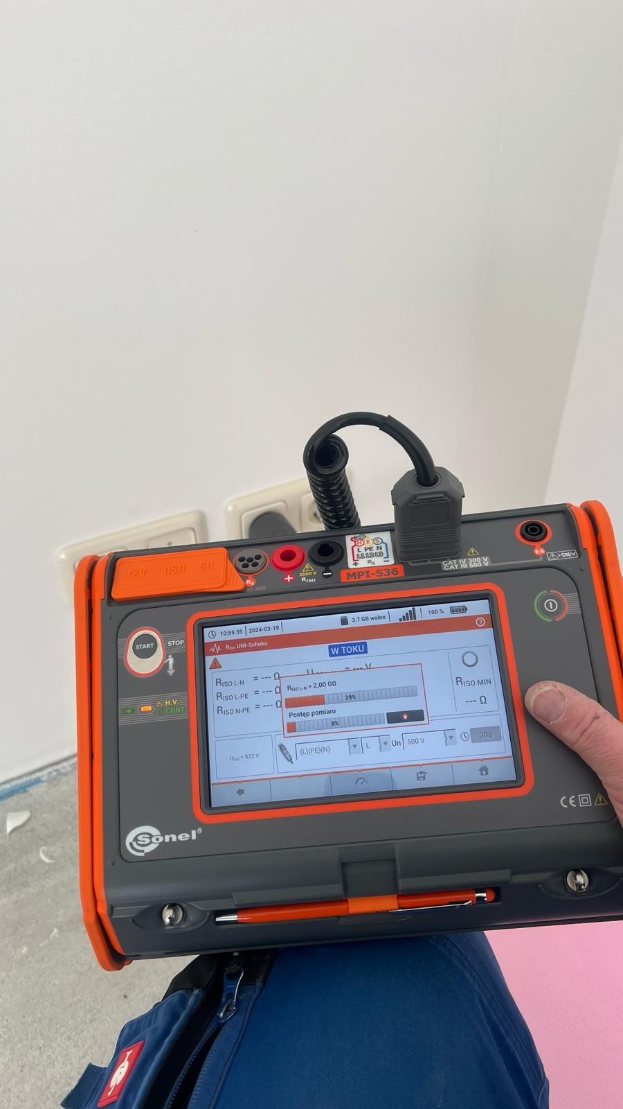 Miernik Sonel MPI-536 podłączony do gniazdka elektrycznego podczas wykonywania pomiarów rezystancji izolacji, widoczny postęp pomiaru na ekranie urządzenia, w tle biała ściana i fragment podłogi...