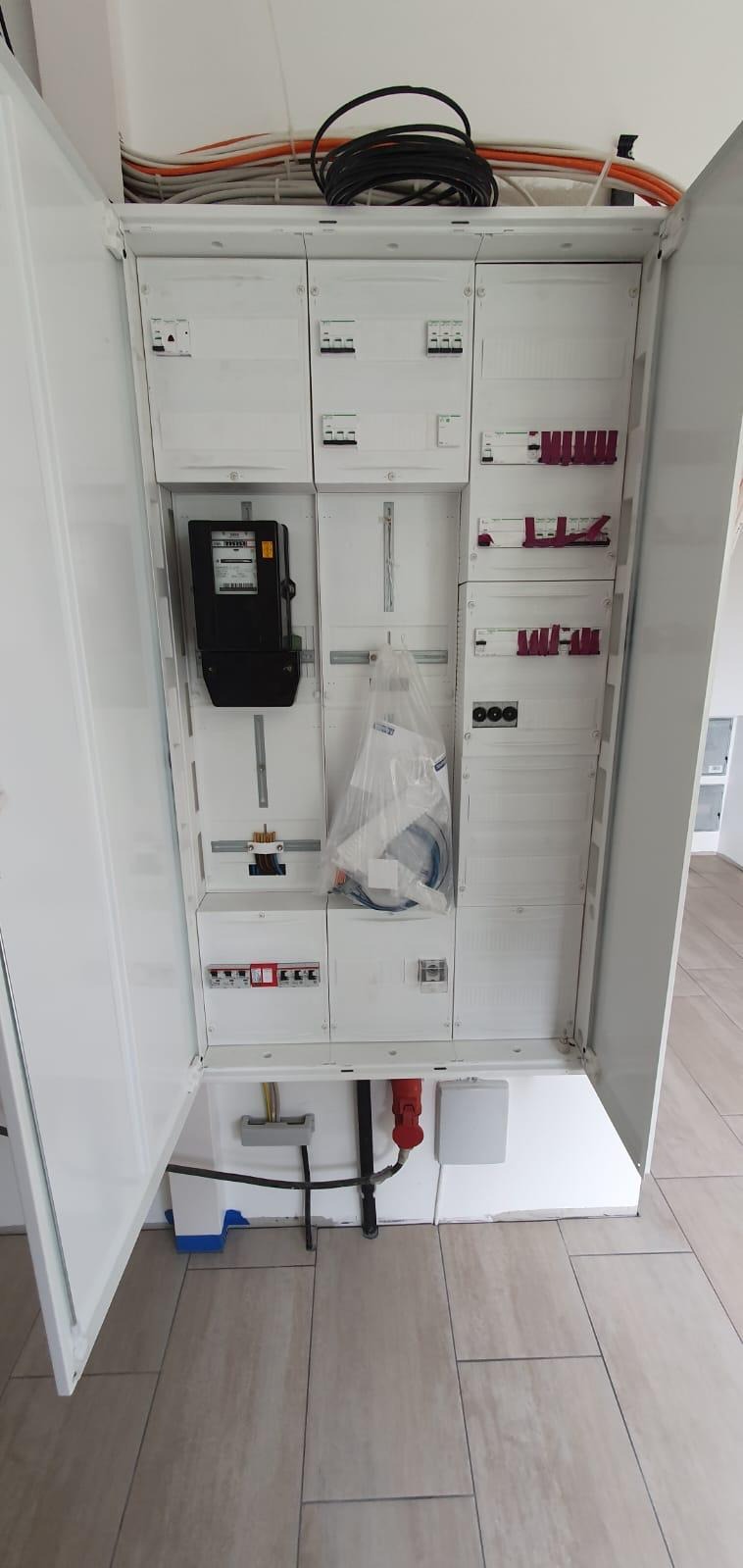 Otwarta, biała szafa rozdzielcza z licznikiem energii elektrycznej i zabezpieczeniami, widoczne kable prowadzone nad szafą i wewnątrz niej, na podłodze płytki imitujące drewno.