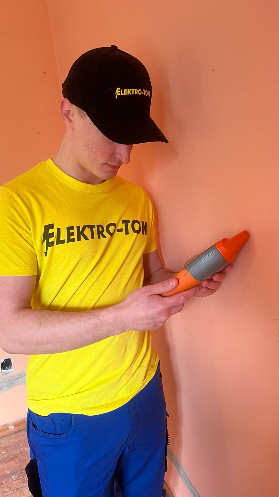 Elektryk w czapce i koszulce z logo firmy Elektro-Tom sprawdza fazę w gniazdku za pomocą próbnika napięcia na tle pomarańczowej ściany.