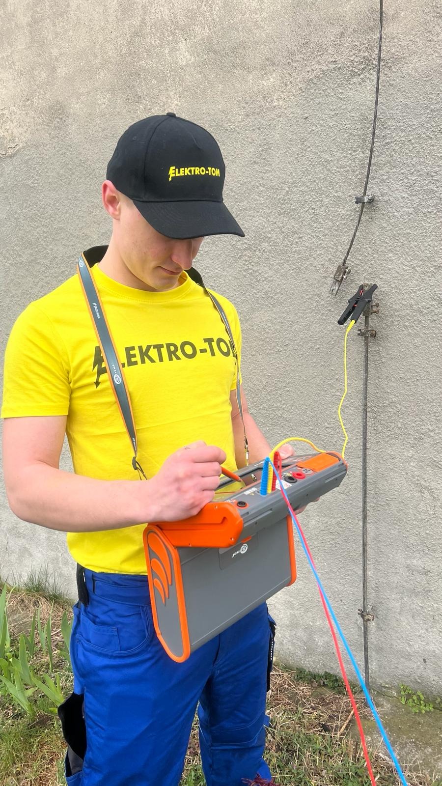 Elektryk w żółtej koszulce i czapce Elektro-Tom wykonuje pomiary uziomu za pomocą miernika, widoczne przewody i uziom przy ścianie budynku.