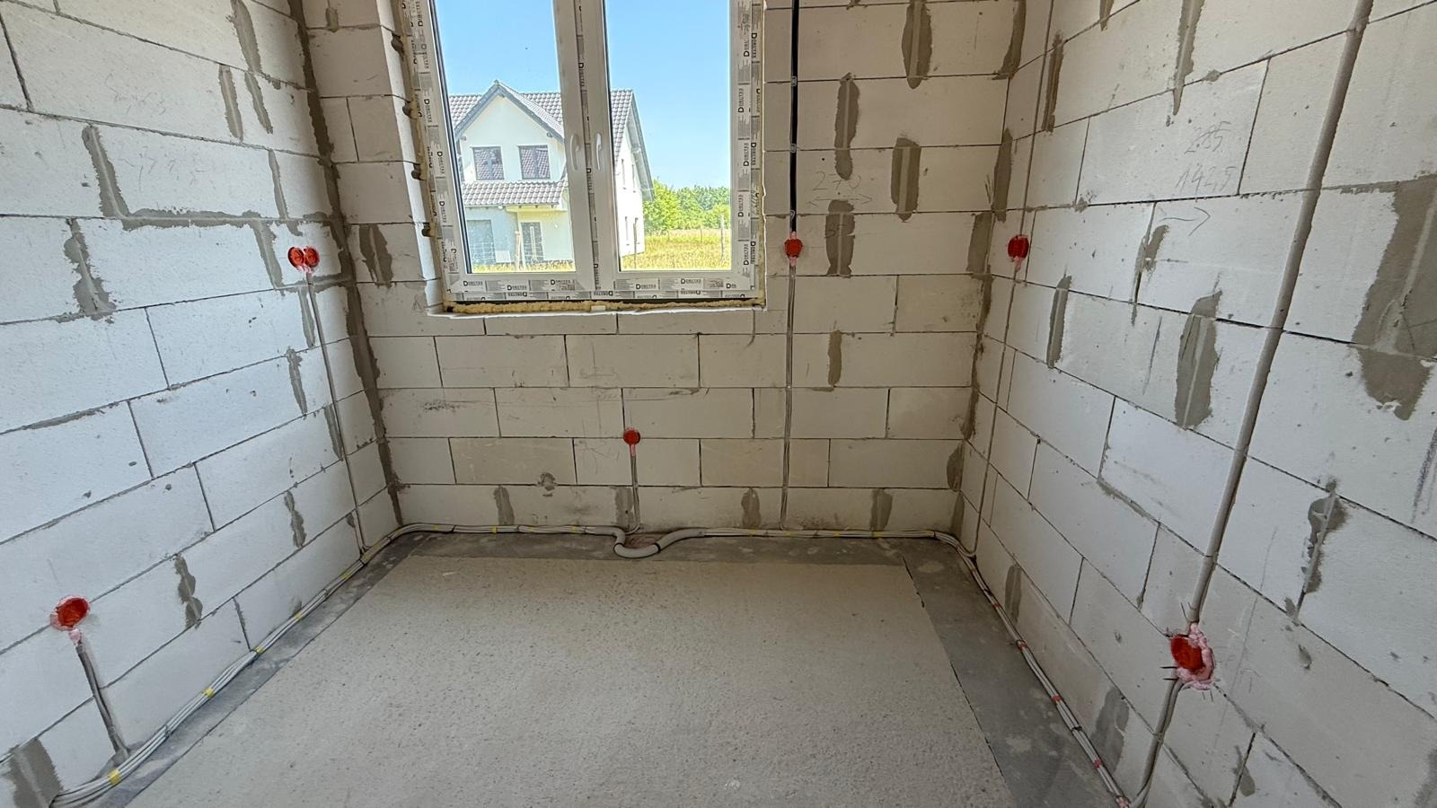 Wnętrze surowego budynku z widoczną instalacją elektryczną: kable wzdłuż ścian, puszki w ścianach z betonu komórkowego. W tle okno z widokiem na inny dom.