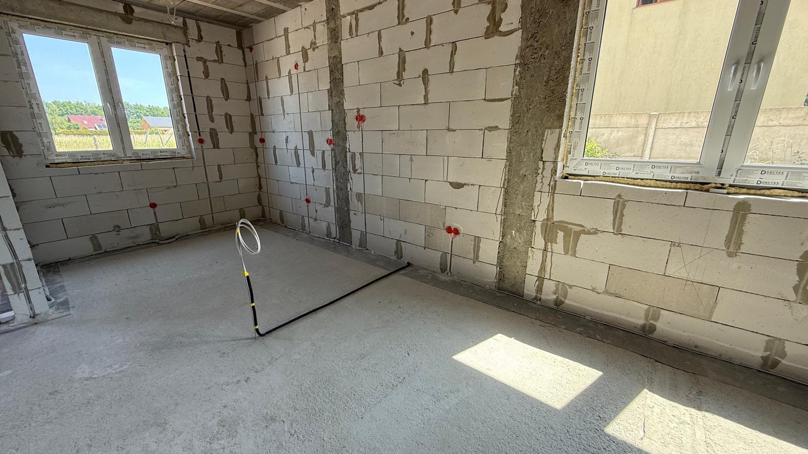 Wnętrze w surowym stanie z widoczną instalacją elektryczną w peszlu, wyprowadzenia kabli w ścianach z betonu komórkowego, okna z widokiem na zieleń.