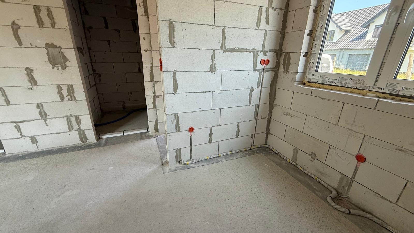 Surowy stan: ściany z betonu komórkowego z puszkami elektrycznymi i przewodami, widoczne okno i sąsiedni budynek. Instalacja w trakcie prac.