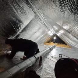 FIRMA OGÓLNOBUDOWLANA SZYMON HALMAN - Prace izolacyjne poddasza z folią aluminiową i montaż rury wentylacyjnej; widoczna osoba w trakcie pracy, narzędzia i oświetlenie punktowe.