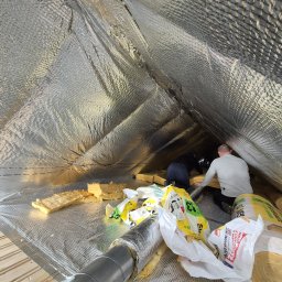 FIRMA OGÓLNOBUDOWLANA SZYMON HALMAN - Dwóch mężczyzn w przestrzeni poddasza, instalujących izolację termiczną z folii aluminiowej i wełny mineralnej, widoczne rolki izolacji i rura wentylacyjna, puszka z klejem i nóż monterski w kadrze.