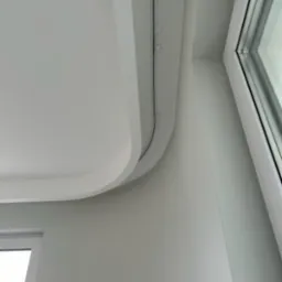 Biały sufit podwieszany z zaokrąglonym narożnikiem i szyną oświetleniową, widok z dołu w rogu pokoju z oknem.