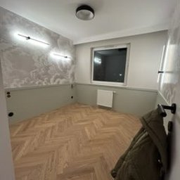 FIRMA OGÓLNOBUDOWLANA SZYMON HALMAN - Pusty pokój po remoncie z nową podłogą w jodełkę, ozdobną tapetą z motywem chmur, oświetleniem LED i grzejnikiem pod oknem, widziany z perspektywy wejścia.