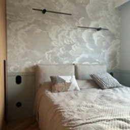 FIRMA OGÓLNOBUDOWLANA SZYMON HALMAN - Sypialnia po remoncie: łóżko z beżowym zagłówkiem i narzutą, ściana z tapetą w motywie chmur, czarne kinkiety, dolna część ściany pomalowana na zielono, widoczny fragment szafy z drewnianymi drzwiami.