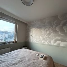 FIRMA OGÓLNOBUDOWLANA SZYMON HALMAN - Sypialnia z tapetą przedstawiającą chmury, pomalowana dolna część ściany na zielono, łóżko z beżową narzutą, widok z okna na budynki.