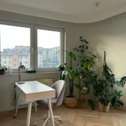 Jasne wnętrze pokoju z parkietem w jodełkę, białym biurkiem z lampką, szarym krzesłem na kółkach i dużymi roślinami doniczkowymi przy ścianie. Widok na miasto za oknem.