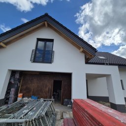 FIRMA OGÓLNOBUDOWLANA SZYMON HALMAN - Dom jednorodzinny w Gdańsku w trakcie prac elewacyjnych. Widoczne rusztowania i materiały budowlane. Biała elewacja, ciemna dachówka i okna.