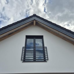 FIRMA OGÓLNOBUDOWLANA SZYMON HALMAN - Biała elewacja domu z czarnym oknem balkonowym i dachem z drewnianymi elementami konstrukcyjnymi. Na szybach okna odbijają się chmury.