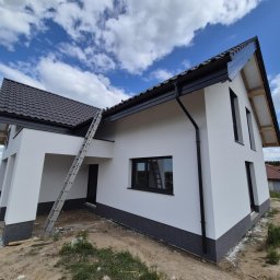 FIRMA OGÓLNOBUDOWLANA SZYMON HALMAN - Nowoczesny dom jednorodzinny z białą elewacją i ciemną podmurówką, drabina oparta o ścianę, błękitne niebo z chmurami w tle. Widok z dołu, podkreślający bryłę budynku.