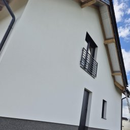 FIRMA OGÓLNOBUDOWLANA SZYMON HALMAN - Biała elewacja domu jednorodzinnego z czarnymi detalami: okna, drzwi, rynna. Widok z dołu, podkreślający nowoczesny styl i dbałość o detale wykończenia.