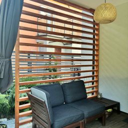 FIRMA OGÓLNOBUDOWLANA SZYMON HALMAN - Drewniana pergola z poziomymi listwami, tworząca intymną przestrzeń na tarasie. W tle widoczny budynek. Na pierwszym planie sofa i lampion.