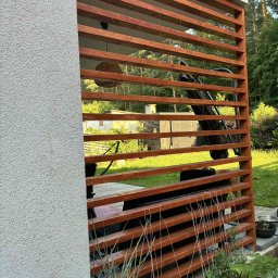 FIRMA OGÓLNOBUDOWLANA SZYMON HALMAN - Drewniana pergola z lamelami, przylegająca do ściany budynku z szarym tynkiem strukturalnym. Widoczne rośliny i elementy wyposażenia tarasu. Ujęcie z bliska.