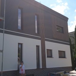 FIRMA OGÓLNOBUDOWLANA SZYMON HALMAN - Dwukondygnacyjny dom z elewacją w kolorach bieli i grafitu, z elementami drewnianymi. Część elewacji w trakcie prac, widoczne rusztowania. Słoneczny dzień.