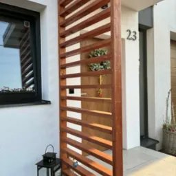 Drewniana, ażurowa przesłona tarasowa z numerem domu 23, zamontowana przy białej elewacji. Dekoracyjny lampion i kwiaty w oknie dodają uroku.