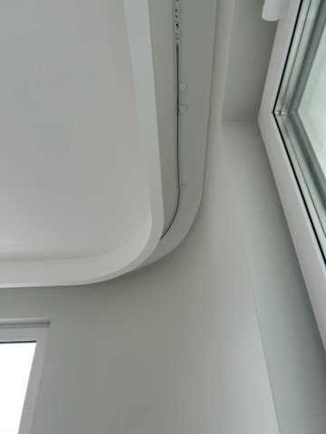 Biały sufit podwieszany z zaokrąglonym narożnikiem i szyną oświetleniową, widok z dołu w rogu pokoju z oknem.