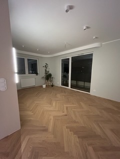 Jasne, przestronne wnętrze z parkietem ułożonym w jodełkę, widok na balkon przez duże przesuwne okno, minimalistyczne oświetlenie sufitowe i roślina doniczkowa przy oknie.