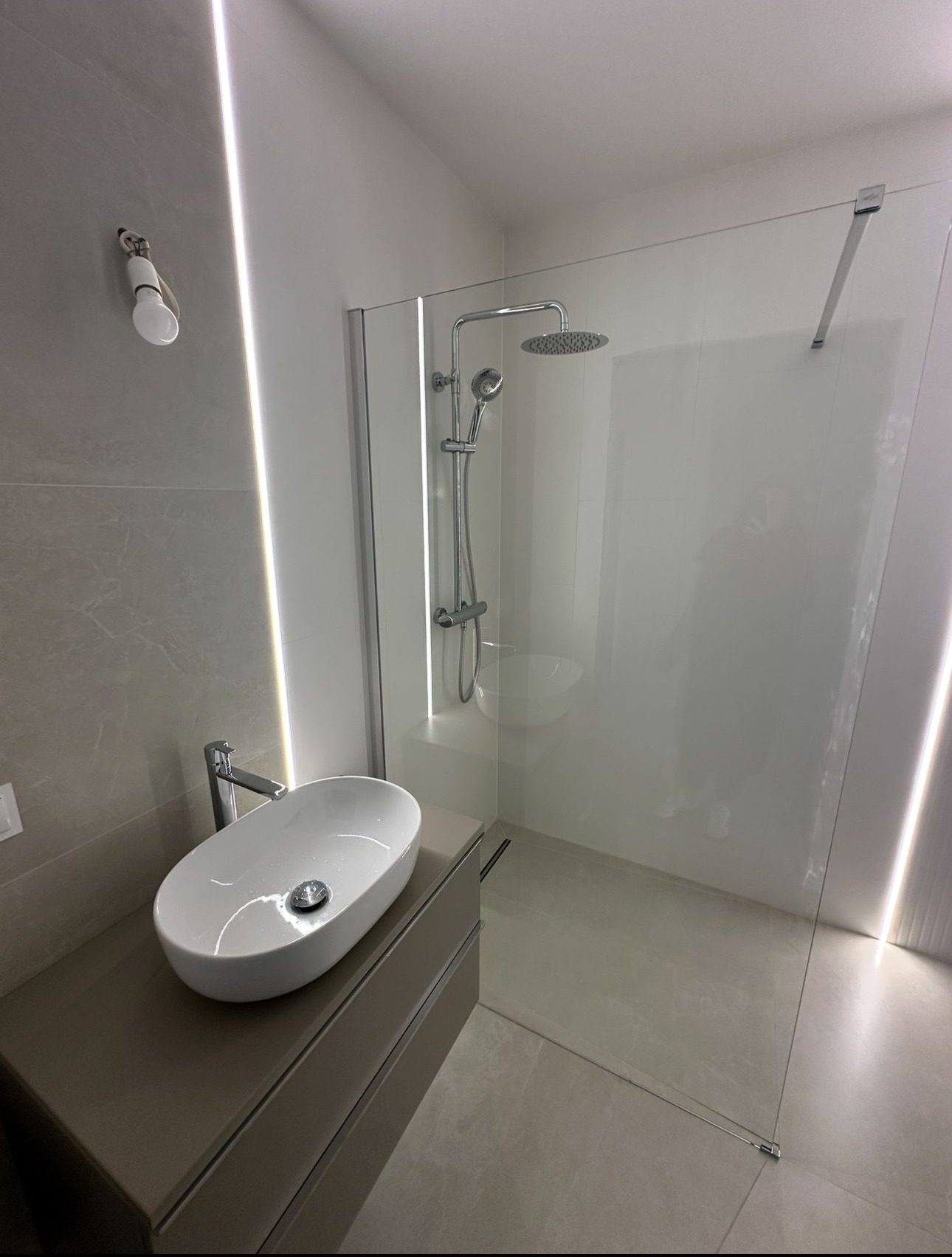 Nowoczesna łazienka z kabiną typu walk-in, owalną umywalką nablatową i subtelnym oświetleniem LED. Minimalistyczny design, jasne kolory i eleganckie wykończenie.
