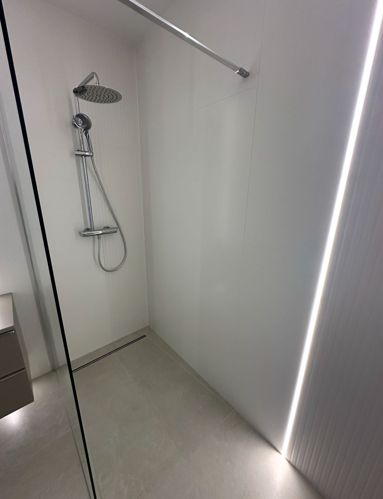 Minimalistyczna kabina prysznicowa z deszczownicą i odpływem liniowym, wykończona w bieli i szarości. Oświetlenie LED wzdłuż ściany dodaje nowoczesnego charakteru.