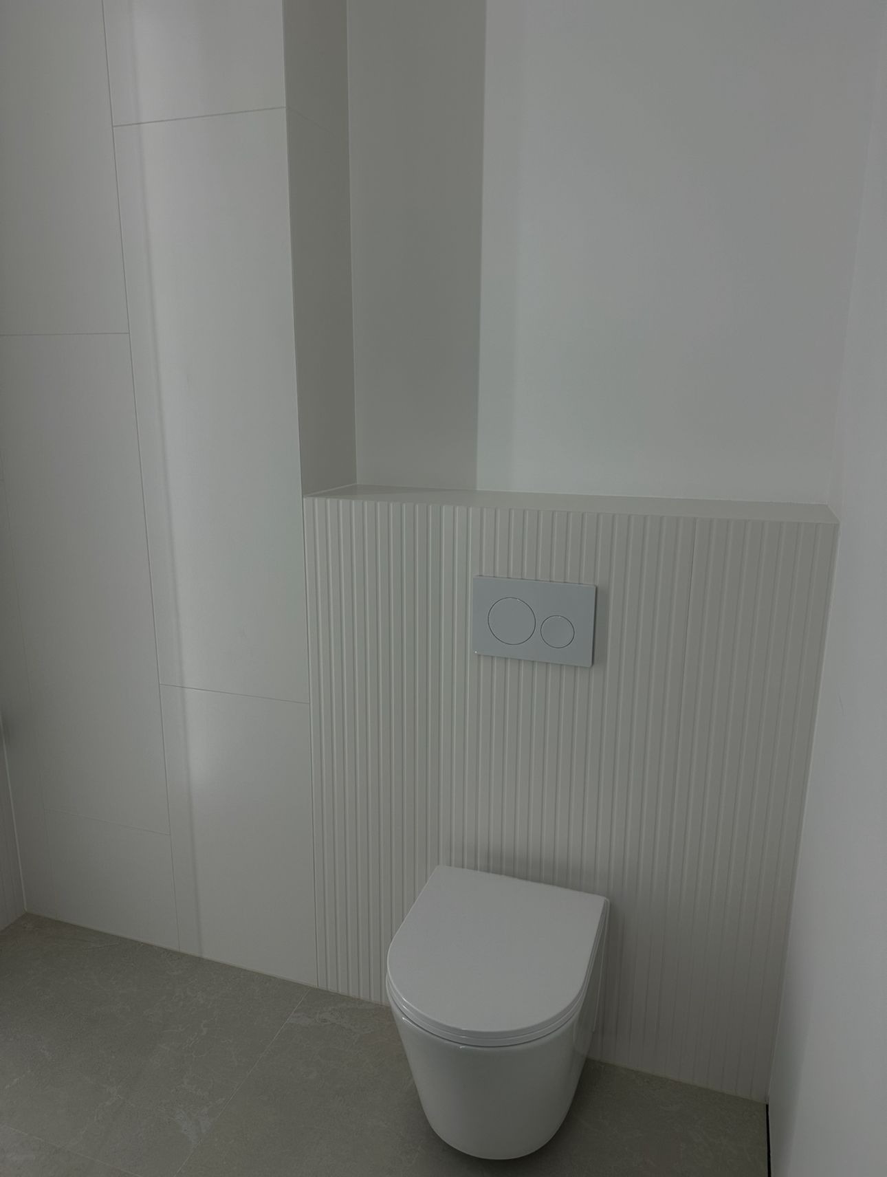 Nowoczesna, minimalistyczna toaleta w odcieniach bieli z subtelną, pionową fakturą na ścianie i szarą podłogą. Widoczny przycisk spłukiwania.