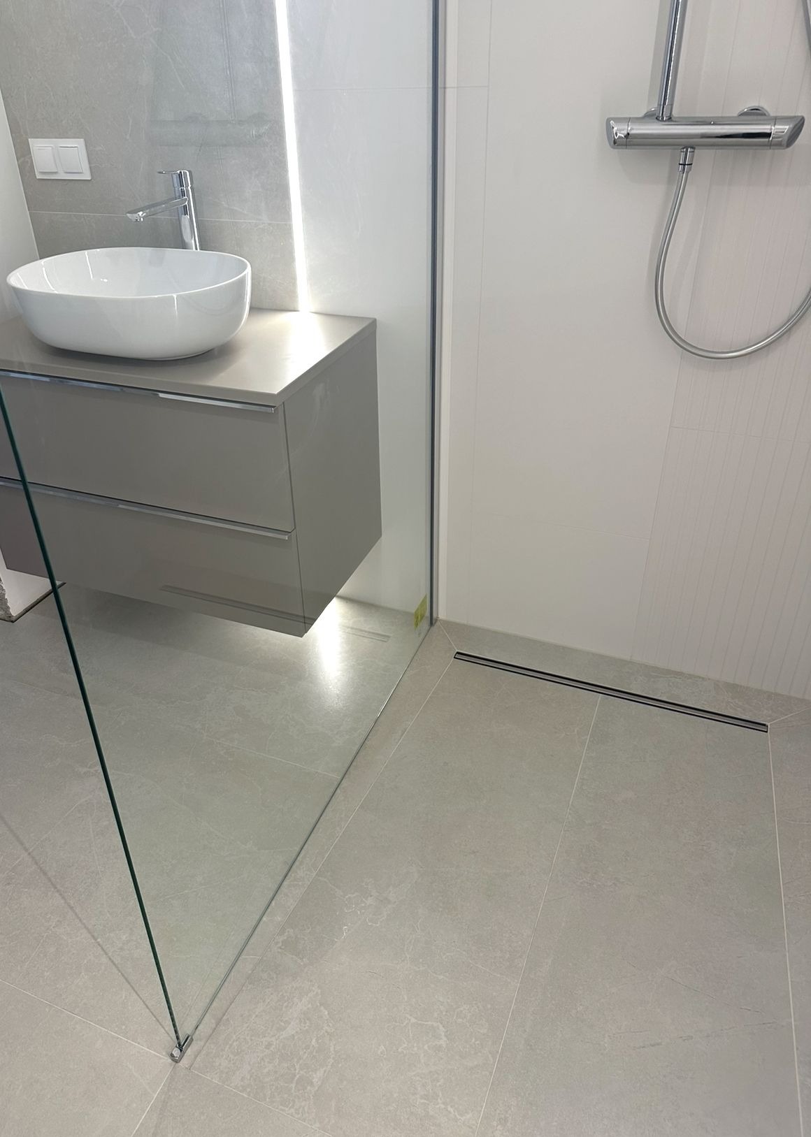 Nowoczesna łazienka z umywalką nablatową, szklaną kabiną prysznicową i odpływem liniowym w podłodze. Minimalistyczny design w odcieniach szarości i bieli.