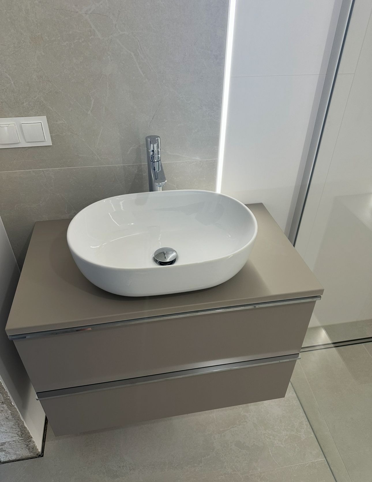 Nowoczesna łazienka: biała, owalna umywalka nablatowa na szafce z szufladami w odcieniach beżu, szara marmurowa ściana i podświetlane lustro.