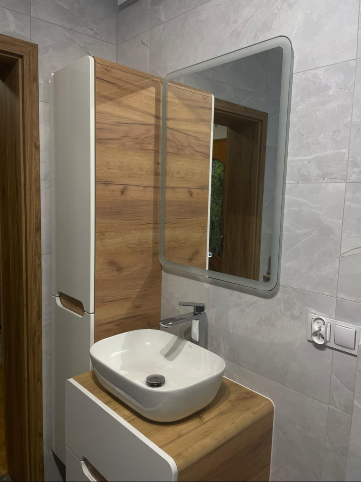 Nowoczesna łazienka: biała umywalka nablatowa, szafka z drewnianym akcentem, lustro z podświetleniem LED i szare płytki imitujące marmur.