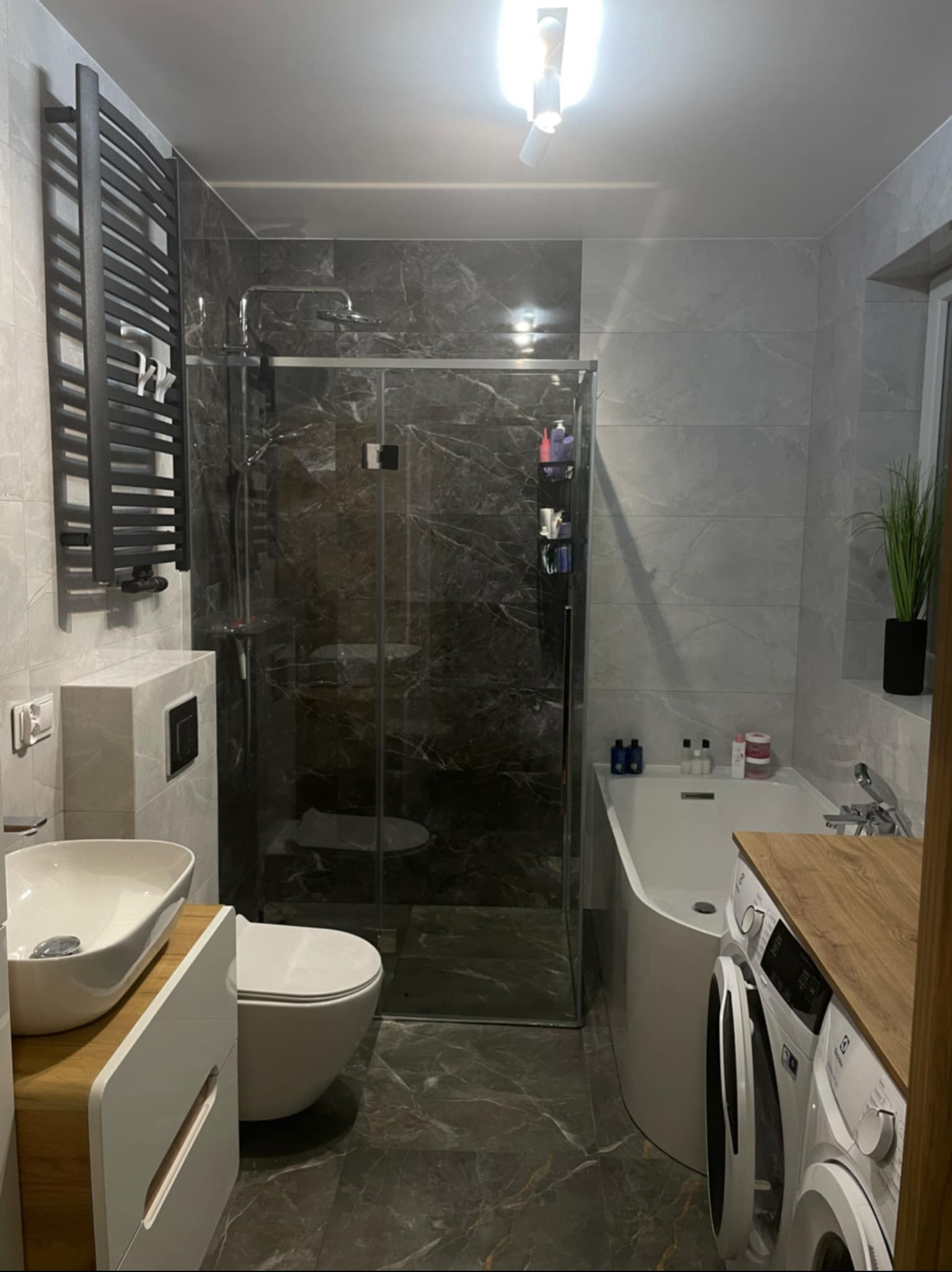 Nowoczesna łazienka z kabiną prysznicową typu walk-in, umywalką nablatową na szafce z drewnianym blatem i grzejnikiem drabinkowym w kolorze czarnym.