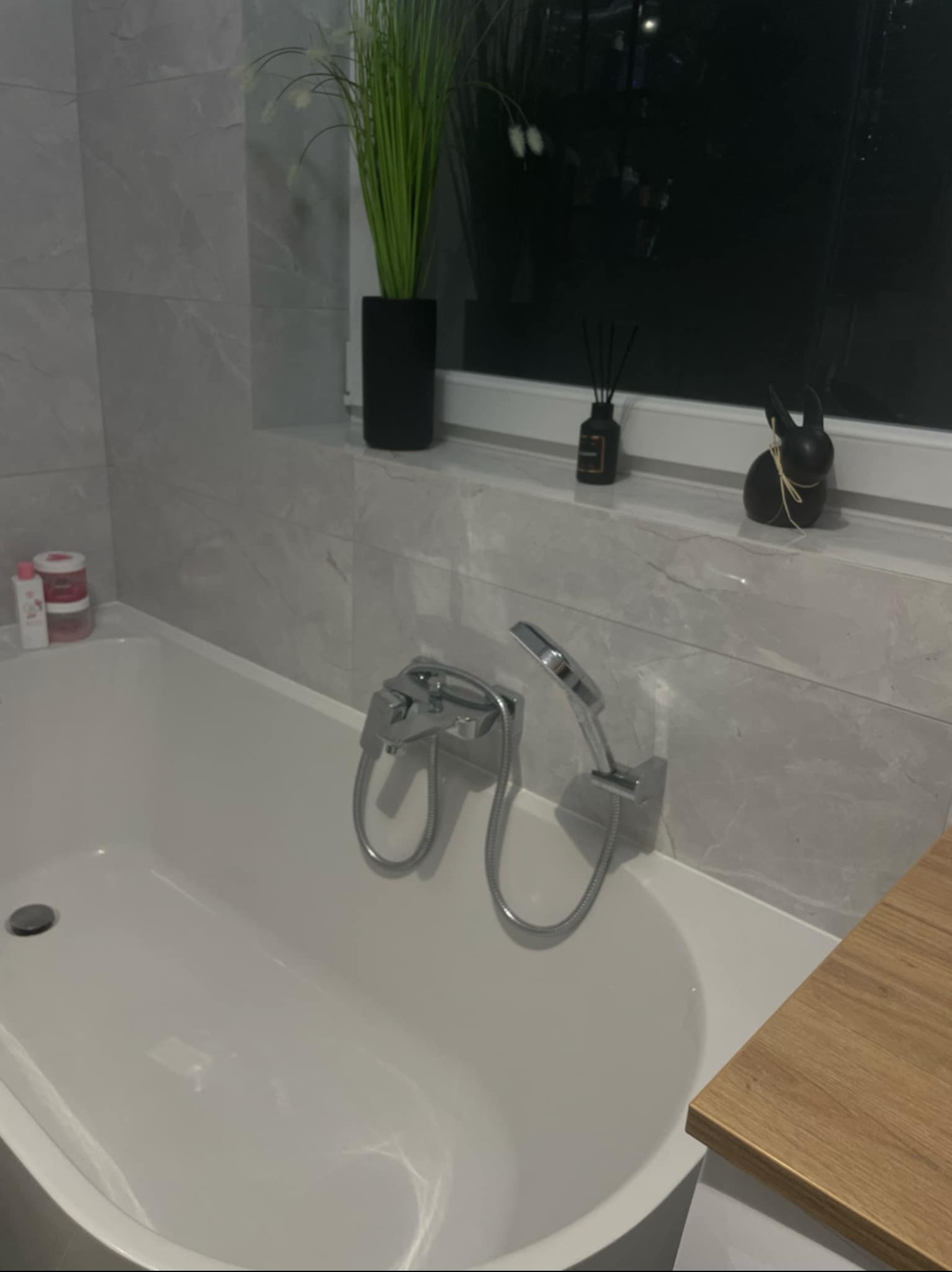 Nowoczesna łazienka z wanną, szarą marmurową okładziną i dekoracjami okiennymi: czarny królik, dyfuzor zapachowy i wysoka roślina w czarnej doniczce.