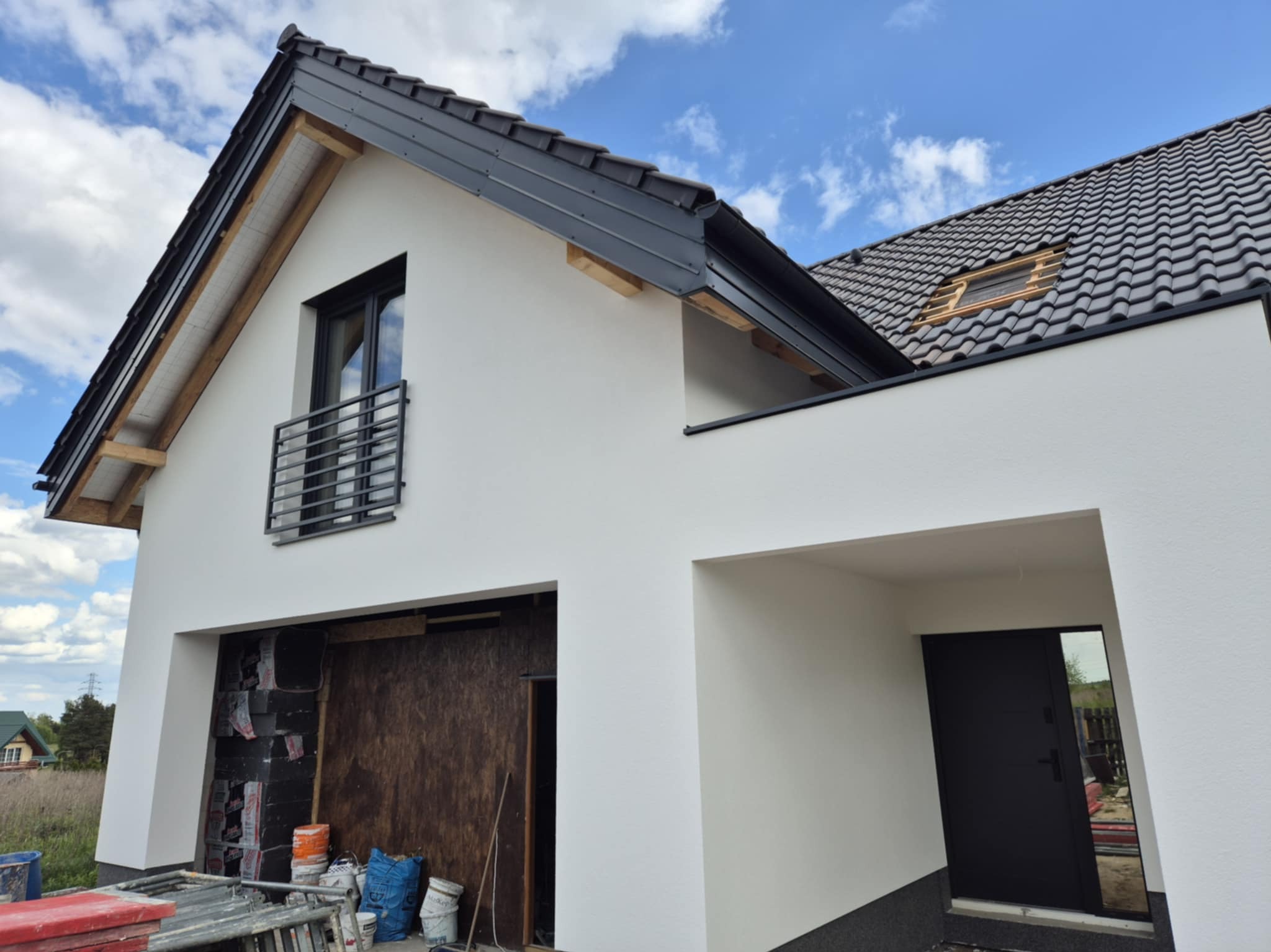 Biała elewacja domu z czarnym dachem i oknami. Częściowo wykończona, widoczne materiały budowlane. Ujęcie z dołu, błękitne niebo w tle.