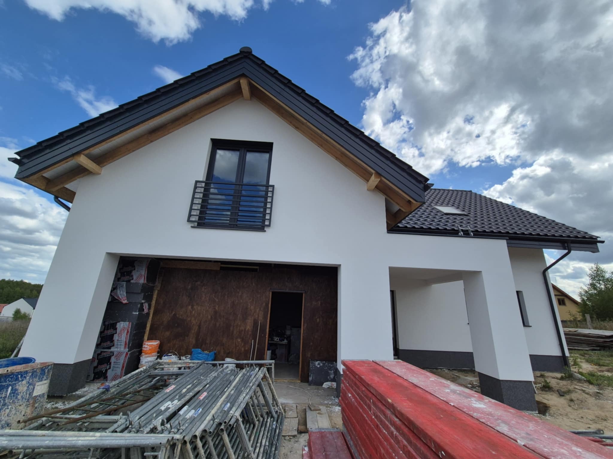 Dom jednorodzinny w Gdańsku w trakcie prac elewacyjnych. Widoczne rusztowania i materiały budowlane. Biała elewacja, ciemna dachówka i okna.