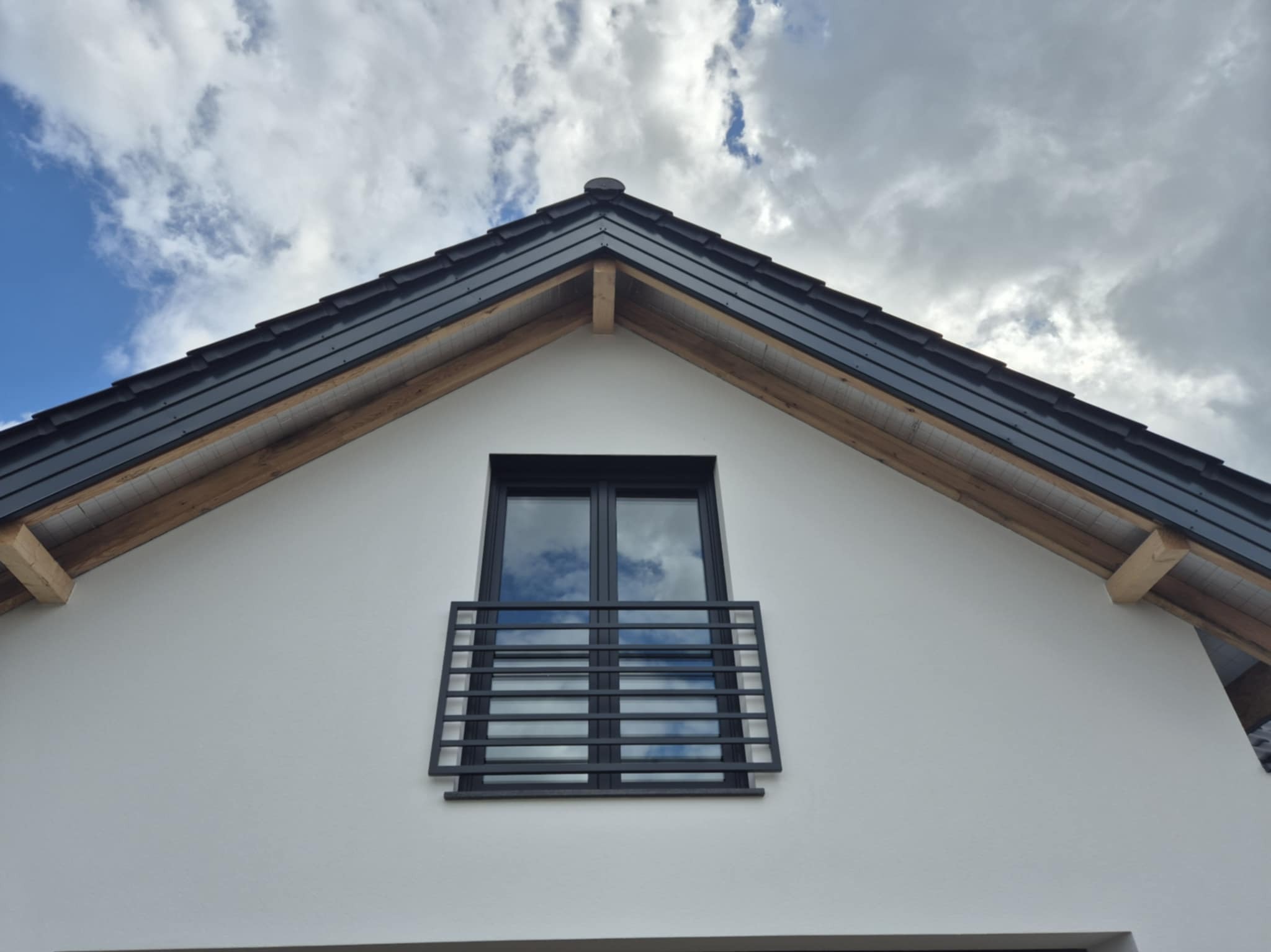 Biała elewacja domu z czarnym oknem balkonowym i dachem z drewnianymi elementami konstrukcyjnymi. Na szybach okna odbijają się chmury.