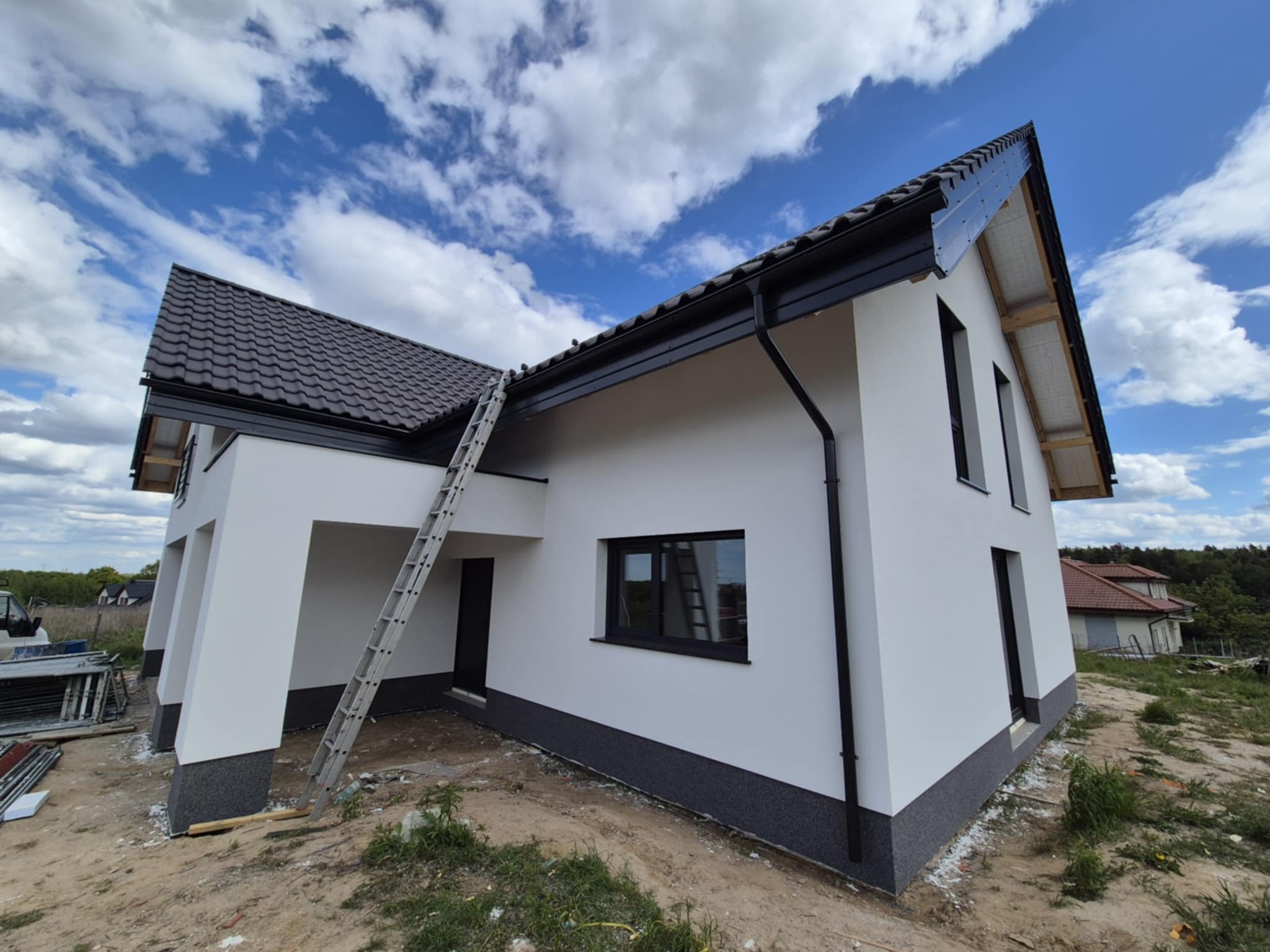 Nowoczesny dom jednorodzinny z białą elewacją i ciemną podmurówką, drabina oparta o ścianę, błękitne niebo z chmurami w tle. Widok z dołu, podkreślający bryłę budynku.