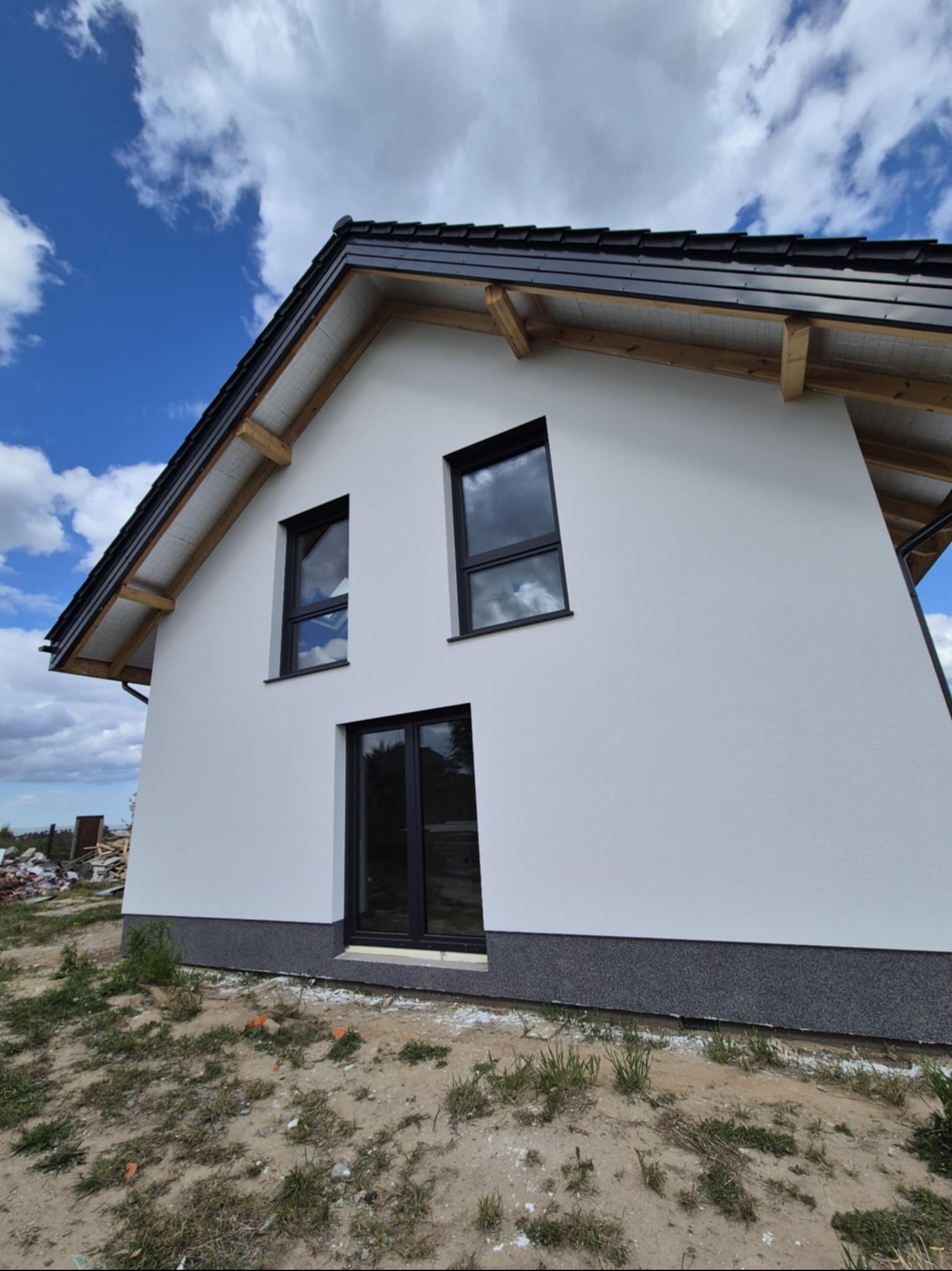 Boczny widok domu z białą elewacją, czarnymi oknami i dachem. Widoczne drewniane elementy konstrukcyjne dachu i fragment niezagospodarowanego terenu.