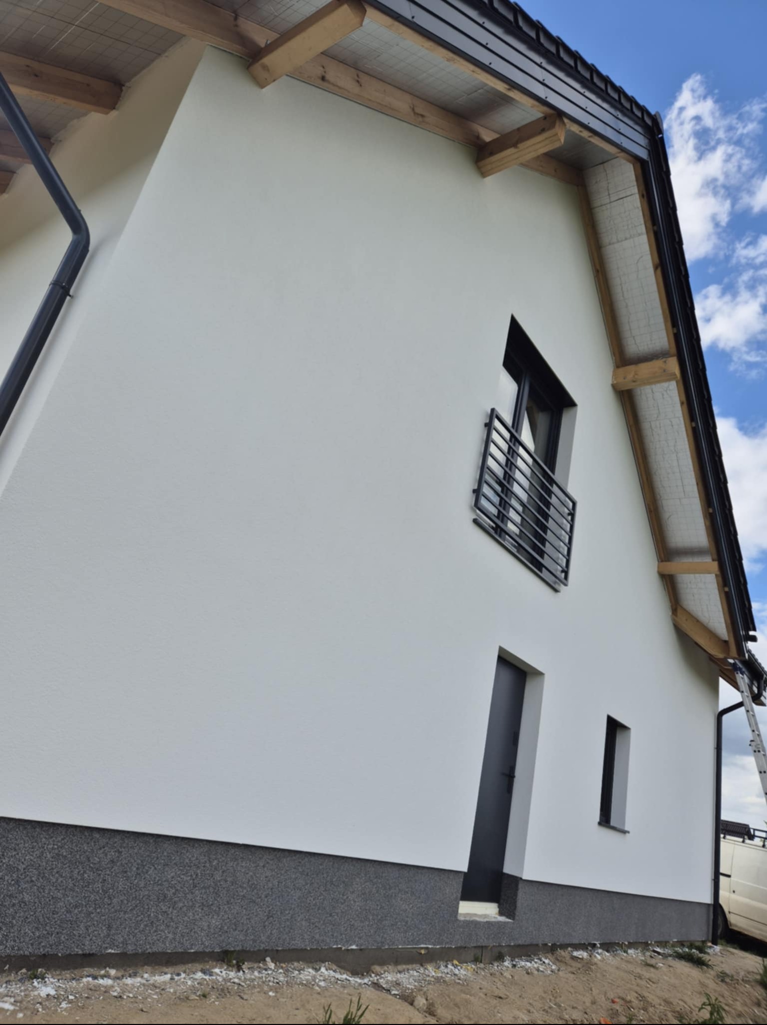 Biała elewacja domu jednorodzinnego z czarnymi detalami: okna, drzwi, rynna. Widok z dołu, podkreślający nowoczesny styl i dbałość o detale wykończenia.