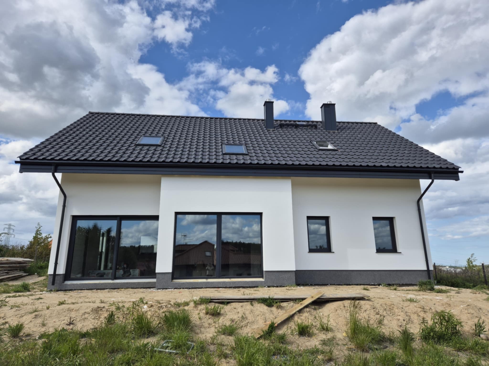 Biały dom jednorodzinny z ciemną dachówką i dużymi oknami, widok z zewnątrz na tle błękitnego nieba z chmurami. Minimalistyczny styl, surowe wykończenie terenu.