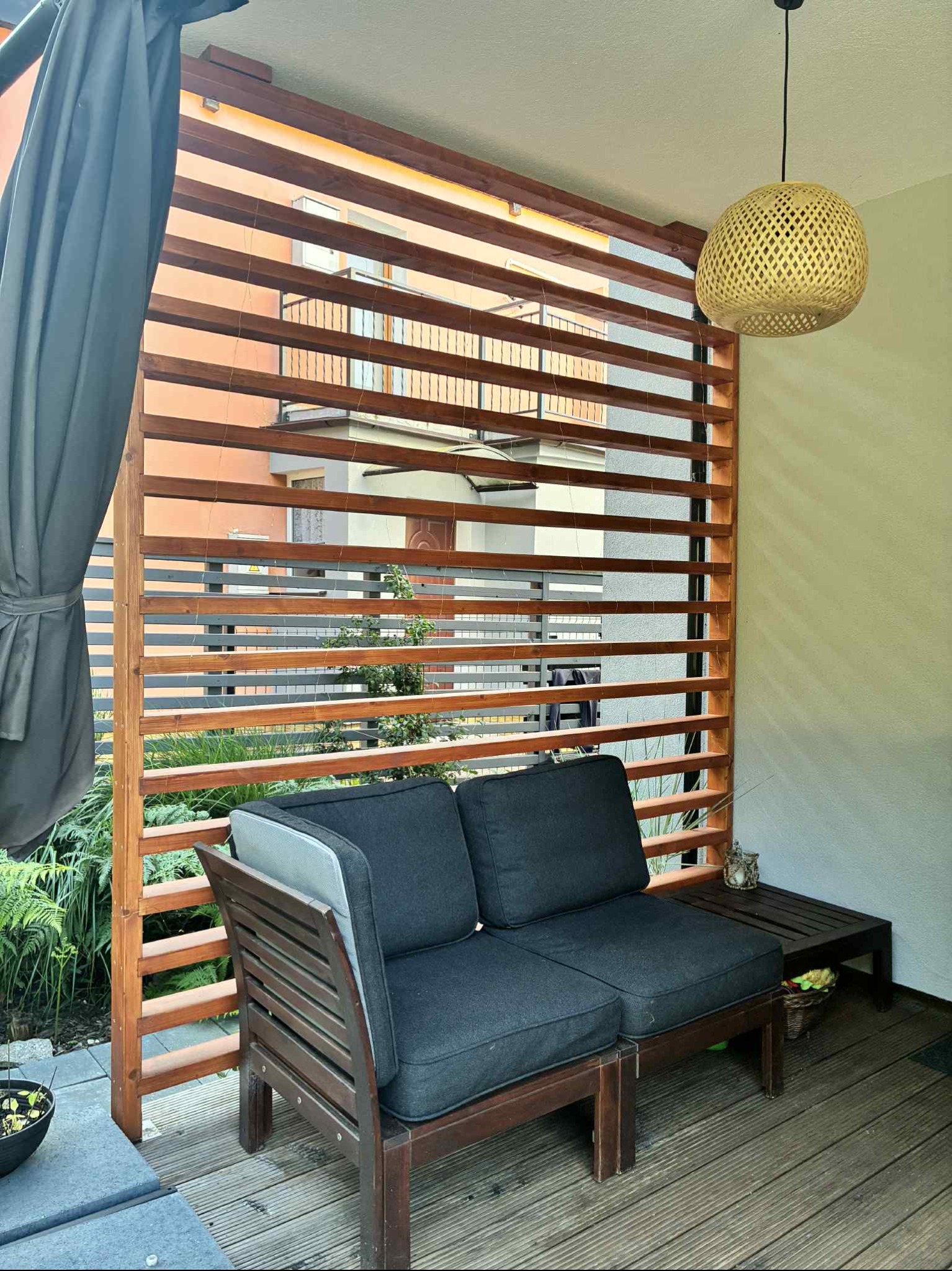 Drewniana pergola z poziomymi listwami, tworząca intymną przestrzeń na tarasie. W tle widoczny budynek. Na pierwszym planie sofa i lampion.