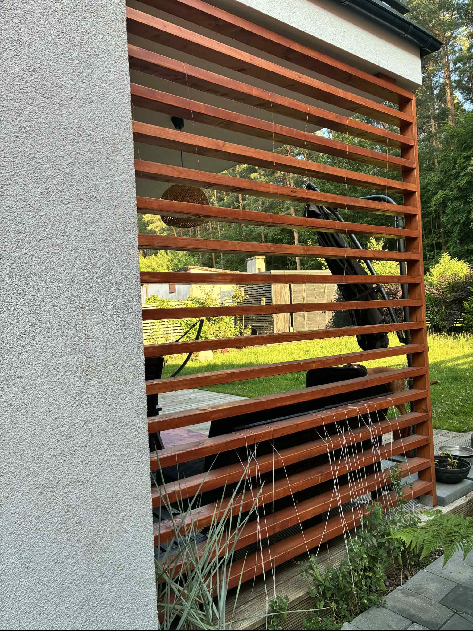 Drewniana pergola z lamelami, przylegająca do ściany budynku z szarym tynkiem strukturalnym. Widoczne rośliny i elementy wyposażenia tarasu. Ujęcie z bliska.