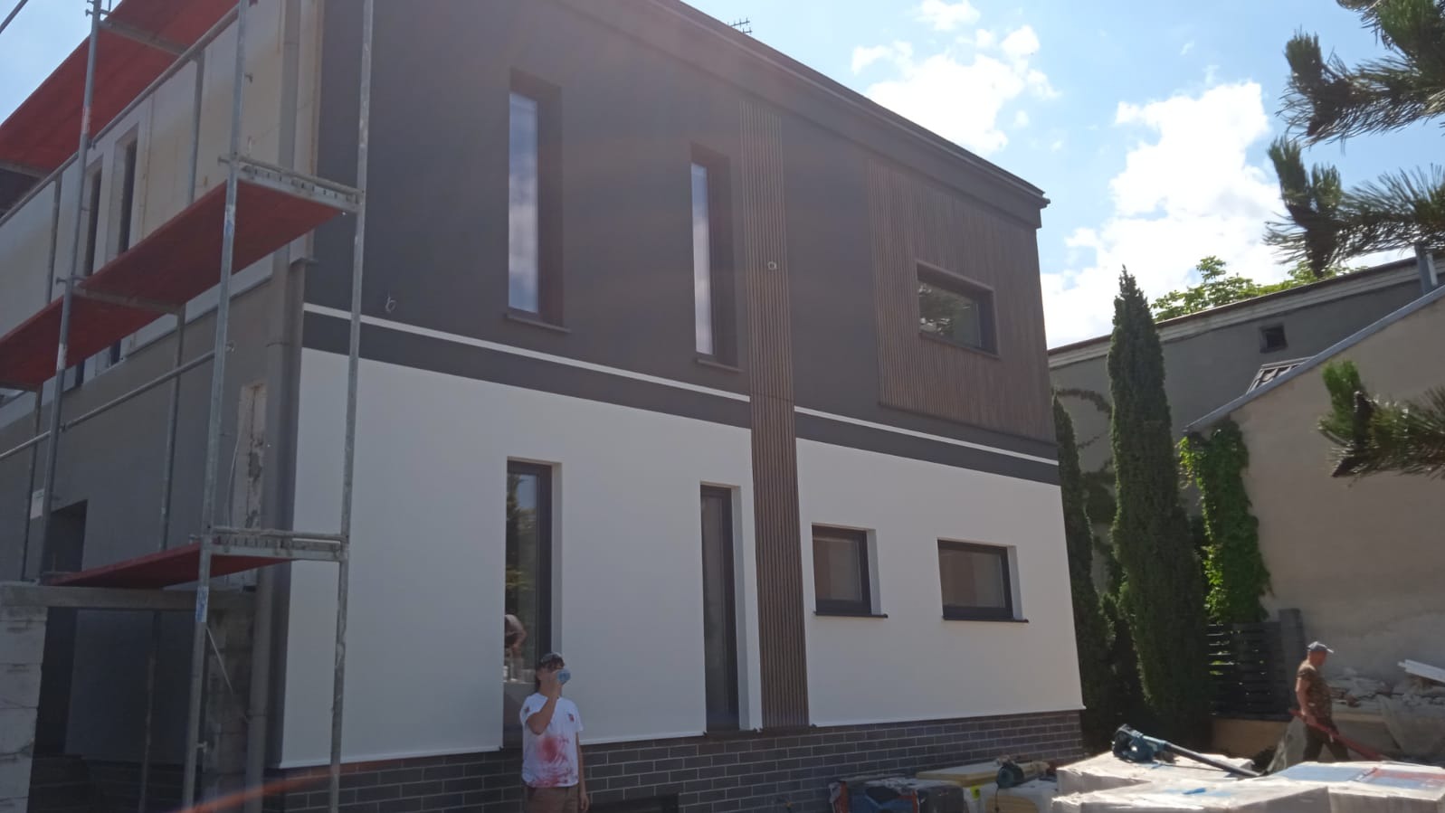 Dwukondygnacyjny dom z elewacją w kolorach bieli i grafitu, z elementami drewnianymi. Część elewacji w trakcie prac, widoczne rusztowania. Słoneczny dzień.