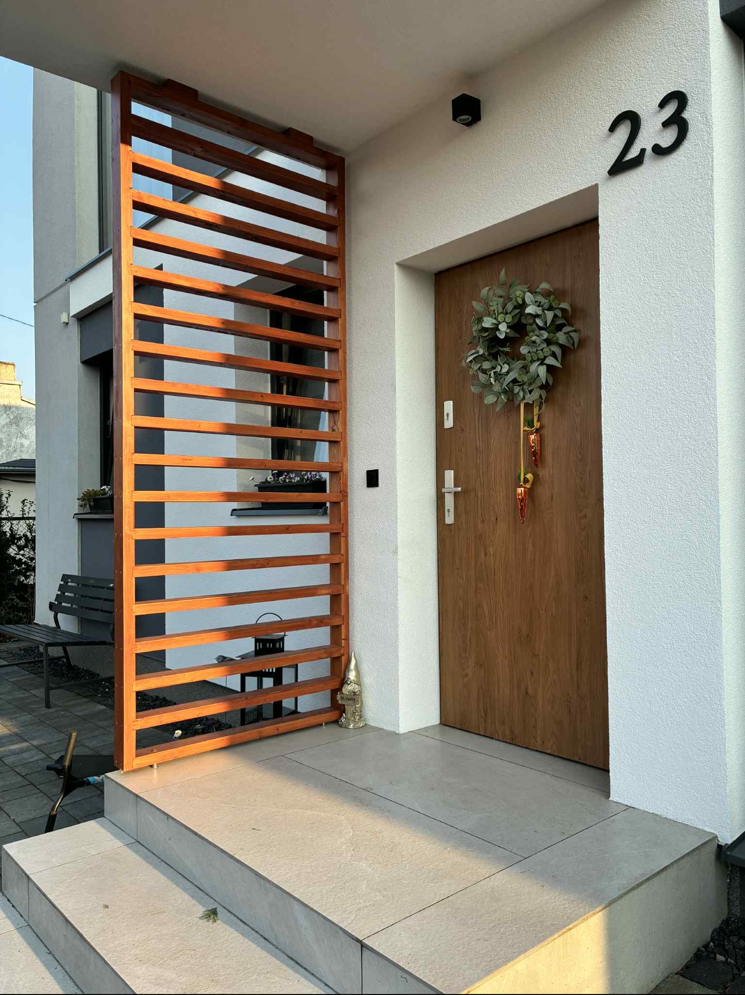 Nowoczesne wejście do domu z drewnianą ażurową przesłoną, numerem 23 nad drzwiami i świątecznym wieńcem. Jasna elewacja i kamienne schody.