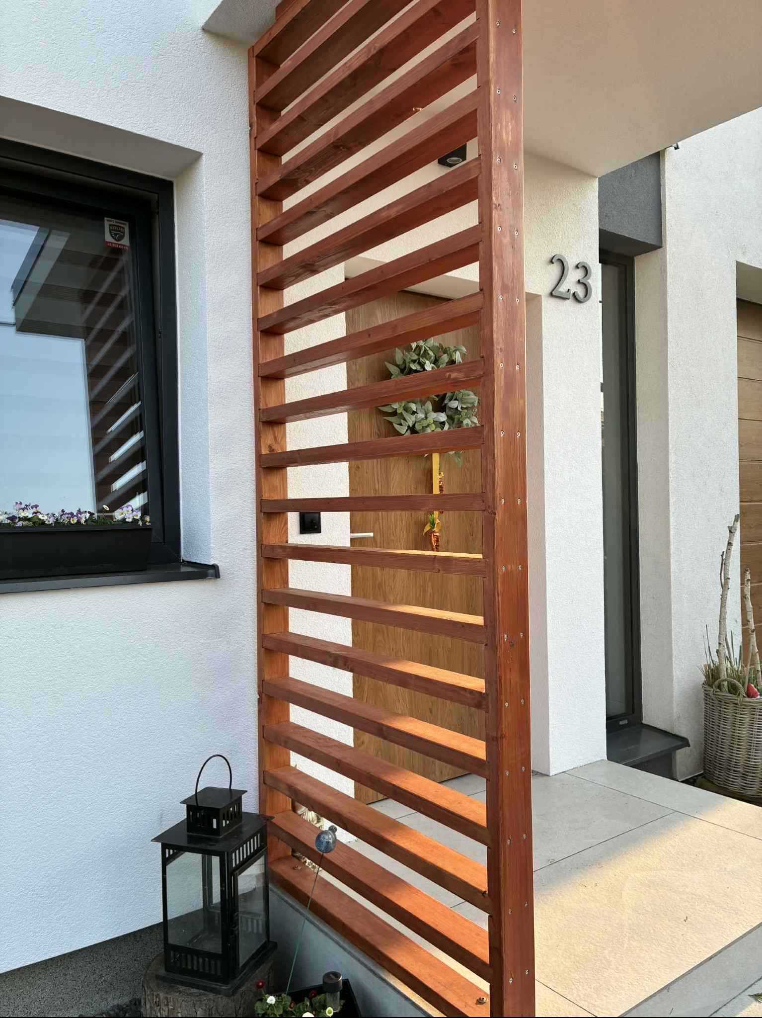 Drewniana, ażurowa przesłona tarasowa z numerem domu 23, zamontowana przy białej elewacji. Dekoracyjny lampion i kwiaty w oknie dodają uroku.