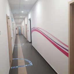 Długi korytarz biurowy z białymi ścianami, szarą podłogą z elementami beżu, drewnianymi drzwiami i dekoracyjnym motywem graficznym w odcieniach różu i fioletu na ścianie. Na podłodze umieszczone są...